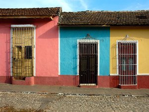 Drie felgekleurde huizen in roze, blauw en geel met pannendaken en deuren met tralies langs een kasseienstraat in Cuba