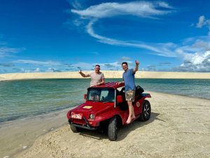 Twee mannen poseren lachend bij een rode buggy op een strand, met een helderblauwe lucht en een lagune op de achtergrond