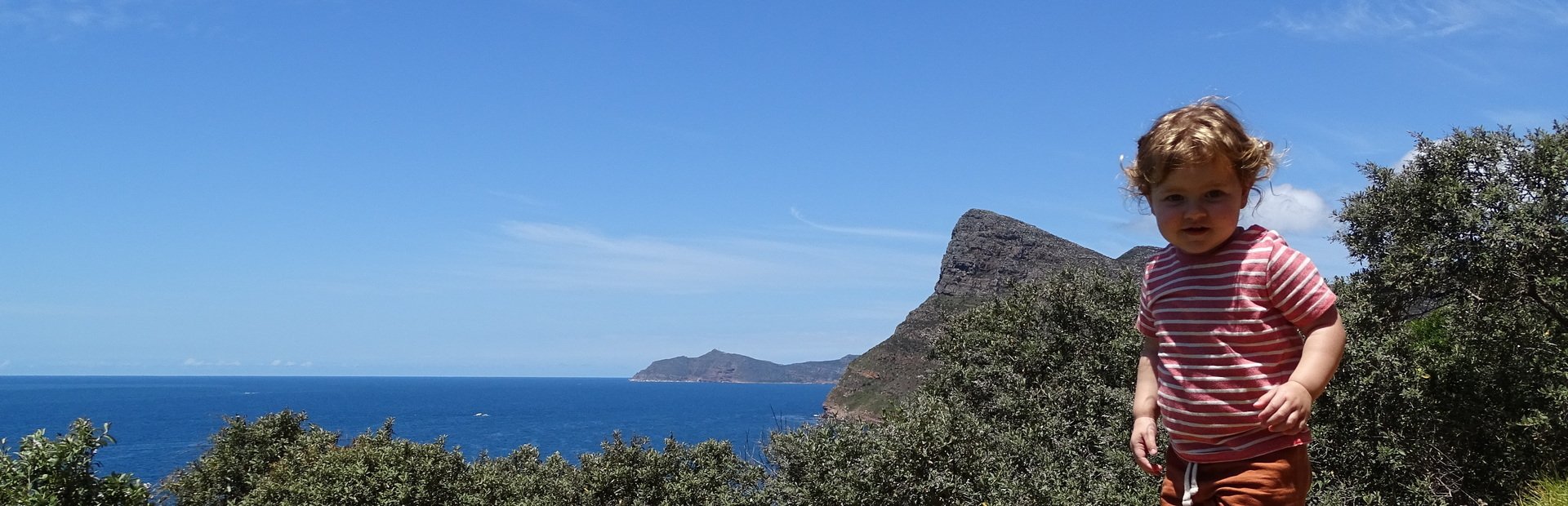 Jong kind staat op de voorgrond met uitzicht op een uitgestrekte blauwe oceaan, bergen in de verte en groene struiken onder een heldere lucht in Zuid-Afrika