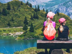 Twee meisjes van achteren gezien zitten op een houten bank en kijken uit over een bergmeer met groene heuvels en bergen in de verte in een natuurgebied