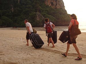 Drie mensen lopen over een strand en trekken elk een koffer achter zich aan. Ze dragen rugzakken en dagrugzakken, op de achtergrond zijn bomen en rotsen te zien.