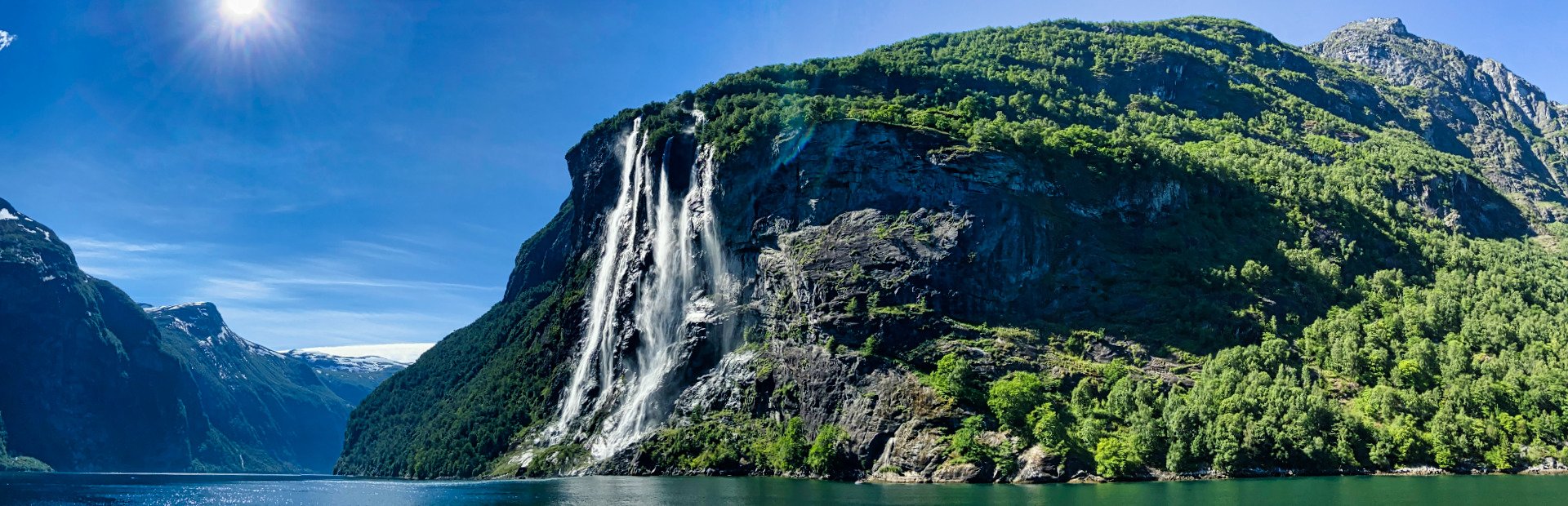 A panoramic view of a fjord with a multi-stream waterfall cascading down a steep, forested cliff under a bright blue sky.