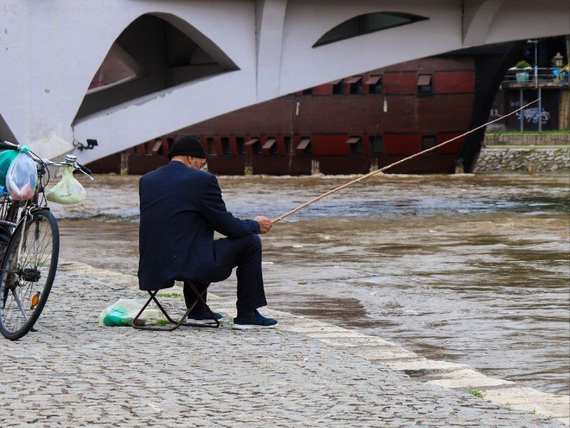 Man in pak vist vanaf een geplaveide rivieroever met een fiets en brug op de achtergrond in Skopje in Noord-Macedonië