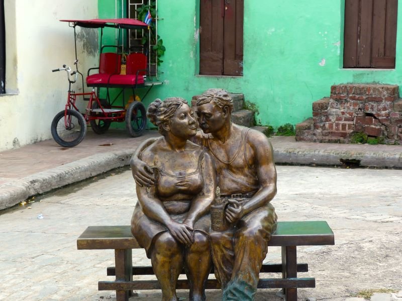 Bronzen standbeeld van een omhelzend stel op een bankje, met op de achtergrond een rode fietstaxi en een groen gebouw in Cuba