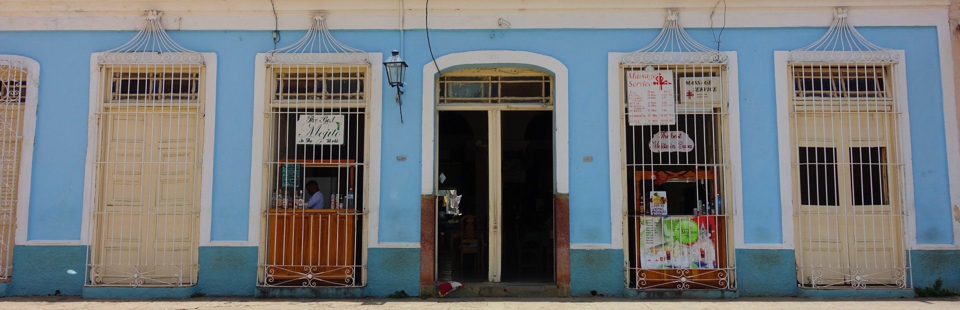 Lichtblauwe gevel met centrale open deuropening en ramen met tralies, waarvan één goederen van een kleine winkel toont in Cuba