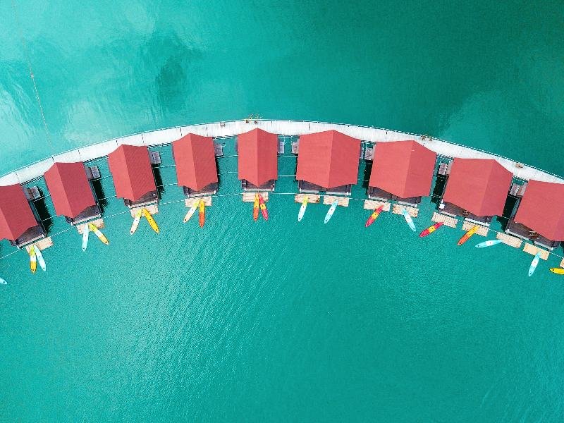 Luchtfoto van een rij drijvende bungalows met rode daken, netjes in een halve cirkel boven turquoise water. Voor elke bungalow liggen kleurrijke kajaks aangemeerd aan een houten vlonder. Het water is kalm en helder.