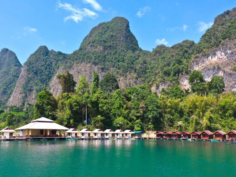 Drijvende bungalows met rieten daken liggen aan een houten steiger op een helderblauw meer, omringd door dichtbegroeide jungle en indrukwekkende, steile kalkstenen rotsen in Khao Sok National Park in Thailand. Op de achtergrond rijzen de groene bergen steil omhoog onder een strakblauwe lucht.
