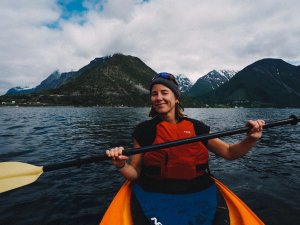 A smiling woman in a red life vest kayaks on a lake with green, snow-capped mountains and a cloudy sky in the background.