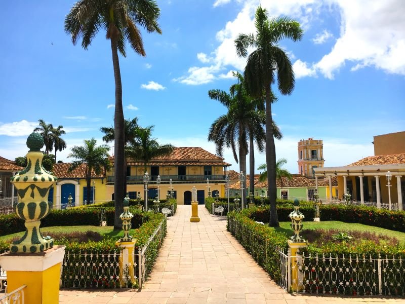 Een historisch park met een geplaveid pad, palmbomen, kleurrijke koloniale gebouwen en een klokkentoren onder een helderblauwe lucht in Cuba