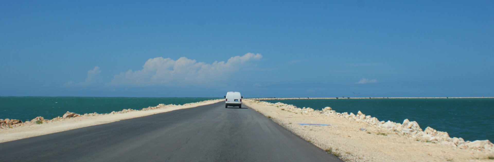 Witte bus rijdt over een weg met aan beide kanten water onder een blauwe lucht in Cuba