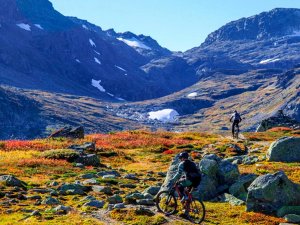 Two mountain bikers on a rocky trail with colorful autumn foliage and mountains.