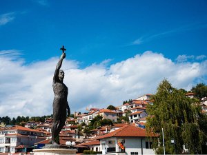 Bronzen standbeeld van een man met een kruis in Ohrid in Noord-Macedonië met uitzicht op een stad met oranje daken op een heuvel onder een blauwe lucht