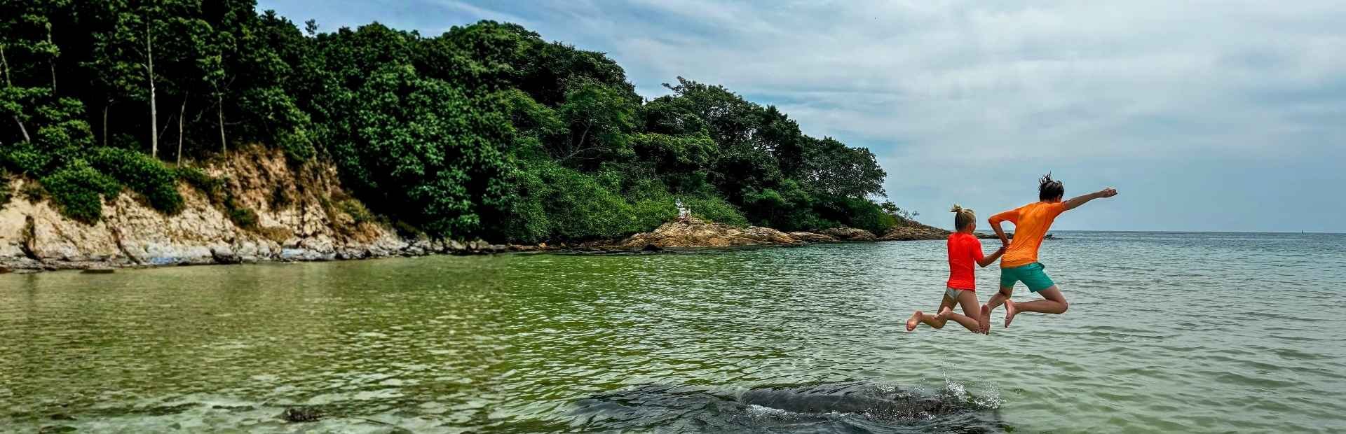 Twee kinderen springen hand in hand het ondiepe zeewater in bij een klein, groen eiland voor de kust van Thailand.
