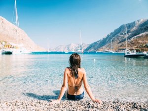 Vrouw in bikini zit op een kiezelstrand met uitzicht op helderblauwe zee, zeilboten en omringende bergen.