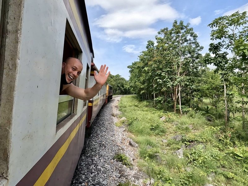 Reiziger steekt lachend zijn hoofd en hand uit het raam van een rijdende trein die door een groen landschap rijdt, met bomen en gras langs het spoor.