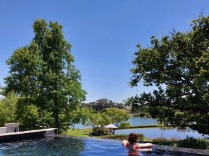Persoon in een infinity pool met uitzicht over een meer omringd door bomen onder een blauwe lucht in Zuid-Afrika