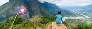 Reiziger zit op bergtop met uitzicht op groene valleien en rivier, naast een wapperende vlag van Laos.