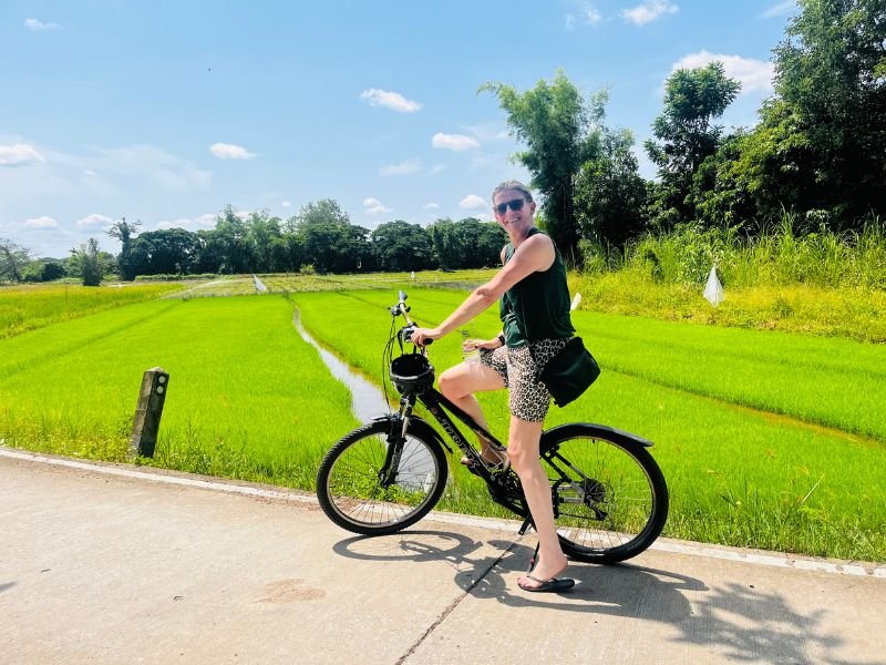 Een vrouw zit op een zwarte fiets op een betonnen weg, met felgroene rijstvelden op de achtergrond. Ze draagt een donkere top, een korte broek met panterprint en een zonnebril. Het is zonnig weer.