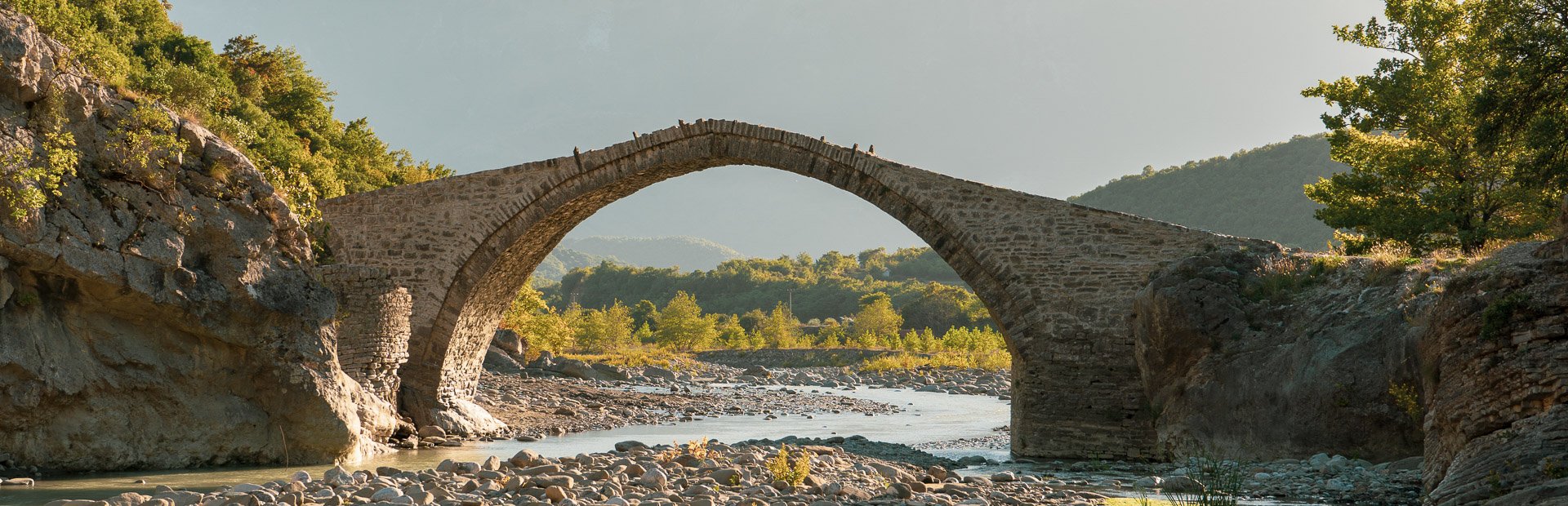 Oude stenen boogbrug in Albanië over een ondiepe rotsachtige rivier met groene heuvels en bomen onder een heldere lucht
