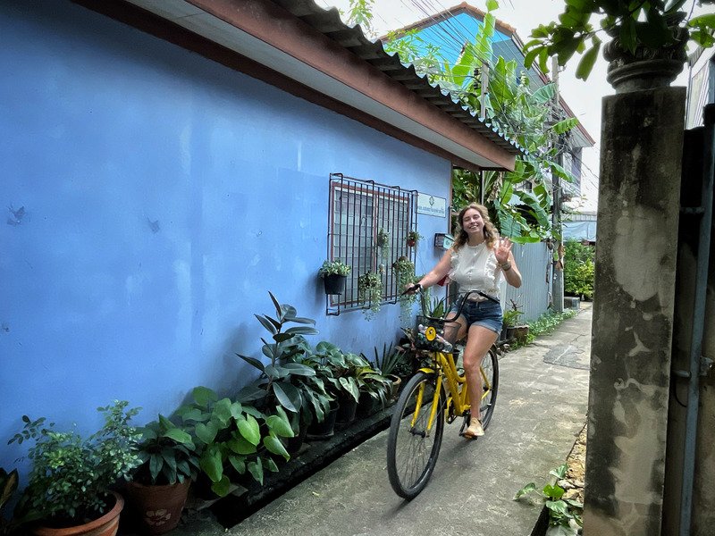Reizigster fietst lachend door een smal straatje in Thailand langs een blauwe muur in Bangkok.