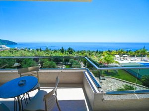 Balcony with a table and chairs overlooking the sea, a beach, green trees, and a resort pool.