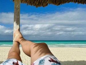 Benen en zanderige voeten rusten onder een rieten parasol, gericht op een tropisch strand met turquoise water.