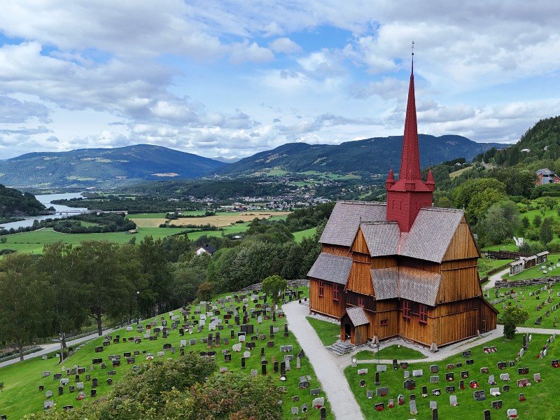A traditional wooden stave church with a red steeple stands in a cemetery overlooking a green valley with a river and mountains.