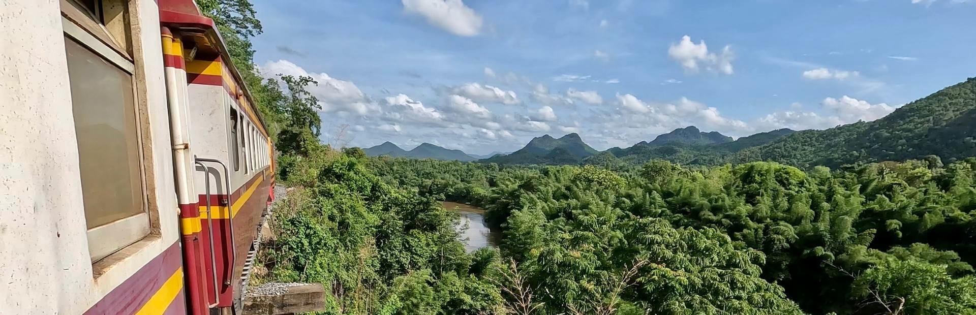 Uitzicht op een beboste vallei met bergen rechts in de verte; links rijdt een rood-wit-gele trein door het landschap. Aan de lucht enkele wolken.