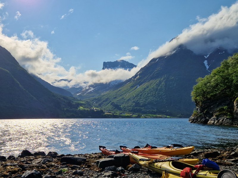 Kayaks on a rocky shore of a sunlit fjord surrounded by majestic, cloud-capped mountains.