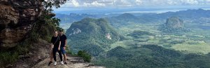 Twee reizigers poseren op een rotsrand met uitzicht op een groen, heuvelachtig landschap en karstbergen in Zuid-Thailand.