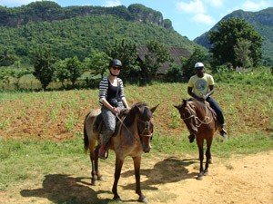 Twee mensen rijden te paard over een zandpad in een landelijke vallei met groene heuvels in Cuba