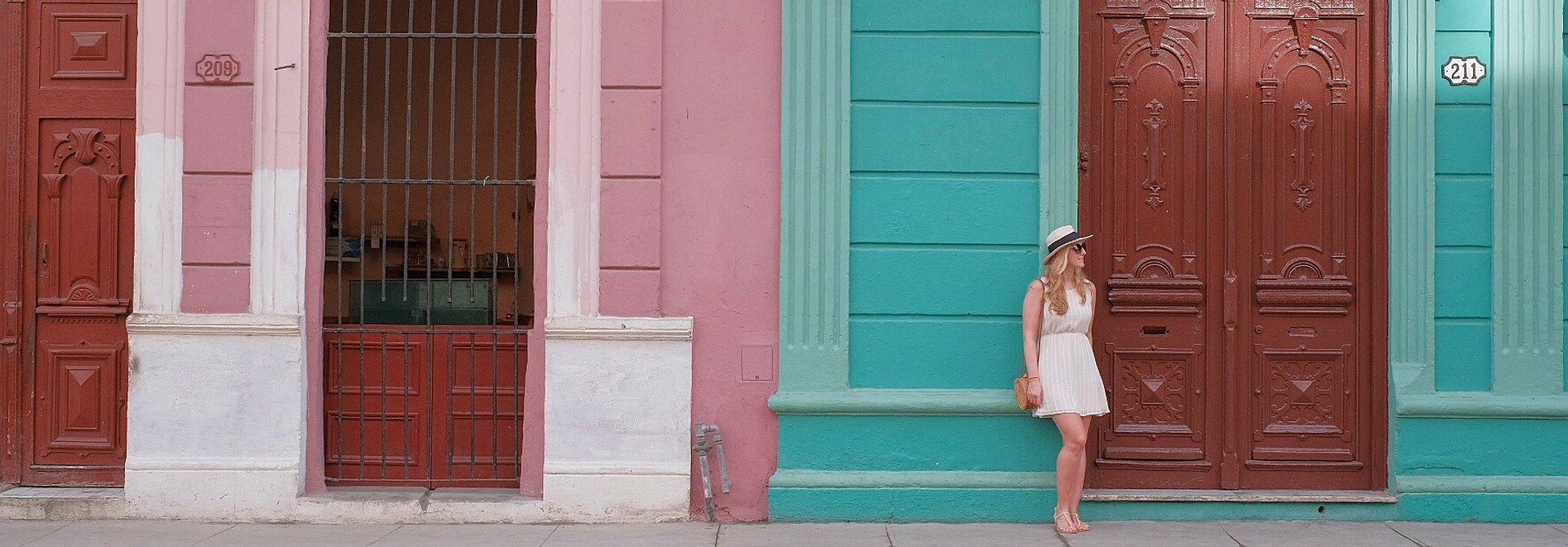 Vrouw in witte jurk en hoed staat naast een grote bruine deur in een teal muur naast een roze gebouw met een tweede deur in Cuba