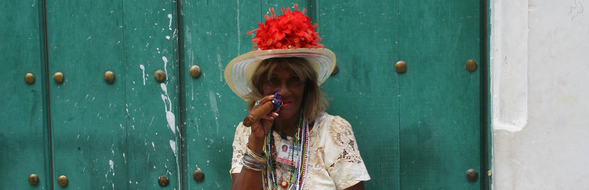 Glimlachende vrouw met bloemenhoed en kralenkettingen houdt een sigaar vast bij een groene deur in Havana Cuba