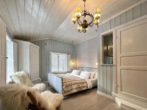 A rustic-style bedroom with light blue walls, a white plank ceiling, a bed with a textured blanket, a white faux fur armchair, and a vintage chandelier.