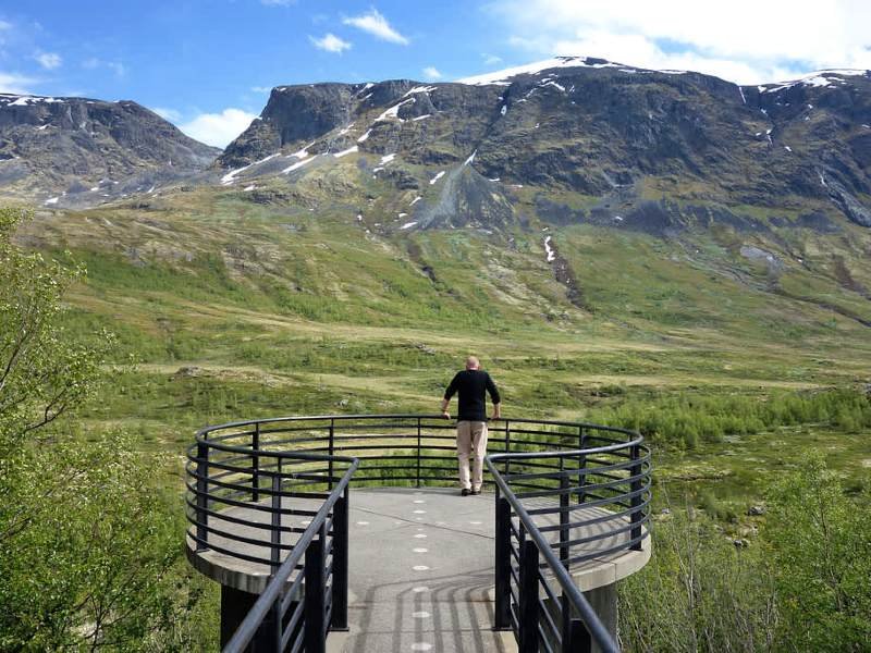 A man on a circular observation deck looks out at a vast green valley surrounded by mountains with snow patches.
