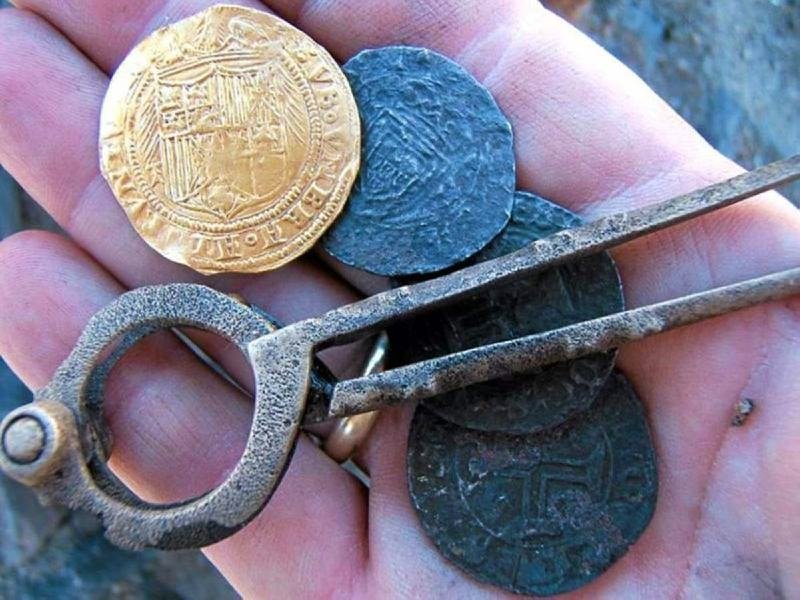 A hand holds one gold coin, several dark coins, and an antique metal coin measuring tool.