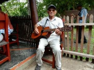 Oudere man in gestreept shirt en pet zit buiten en speelt gitaar in Cuba