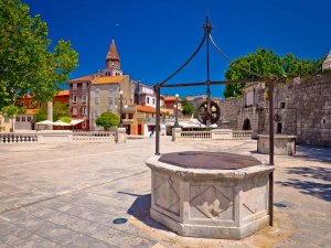 Historische stenen waterput op een plein in Kroatië met kleurrijke gebouwen en een kerktoren onder een heldere blauwe lucht