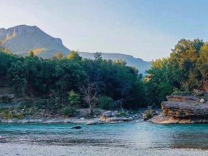 A vibrant turquoise river flows through a valley lined with lush green trees and distant mountains under a clear sky.