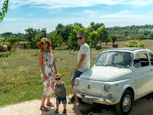 Een vader, moeder en een kindje die staan naast een oude Fiat 500 in Toscane. Ze staan op een zandweggetje, de lucht is blauw met wolken en naast hun is een grasveld met bomen.