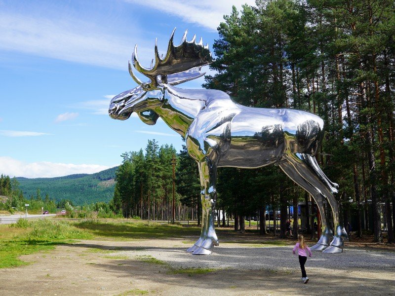 A huge, shiny silver moose sculpture stands in a forest with a child walking near its legs.
