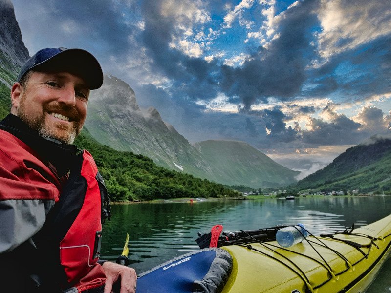Smiling man in a kayak on a lake with mountains and dramatic sky.