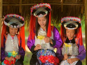 Drie meisjes van een bergstam in Noord-Thailand zitten onder een rieten dak en dragen traditionele, kleurrijke kleding met zilveren details en sierlijke hoofdtooien. Ze zijn geconcentreerd bezig met het maken van bloemenversieringen.