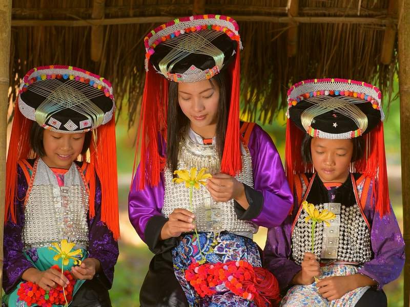 Drie meisjes van een bergstam in Noord-Thailand zitten onder een rieten dak en dragen traditionele, kleurrijke kleding met zilveren details en sierlijke hoofdtooien. Ze zijn geconcentreerd bezig met het maken van bloemenversieringen.