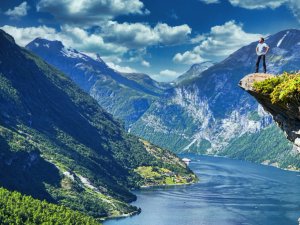 Man standing on a rocky cliff overlooking a vast fjord with steep, green mountains and a cruise ship.