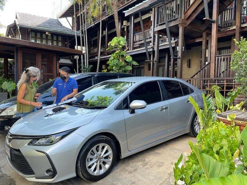 Grijze auto geparkeerd op de oprit van een guesthouse van donker hout met veranda’s en trappen. Naast de auto staat een zwarte auto. Tussen de auto's staan een gids en een vrouwelijke reiziger