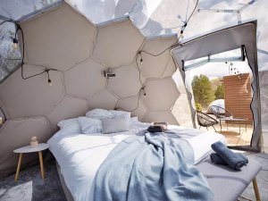 Interieur van een moderne glampingtent met bed, zeshoekige wandpanelen, sfeerverlichting en open doorgang naar een terras in de natuur bij Mavrovo in Noord-Macedonië