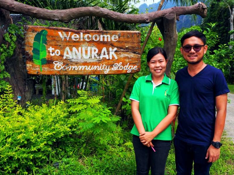 Twee mensen staan voor een houten welkomstbord met de tekst "Welcome to Anurak Community Lodge", omringd door groene planten en een onverhard pad.