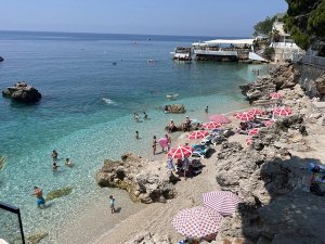 Druk rotsstrand in Albanië met helder turquoise water, rood-witte parasols en een gebouw op de klif op een zonnige dag