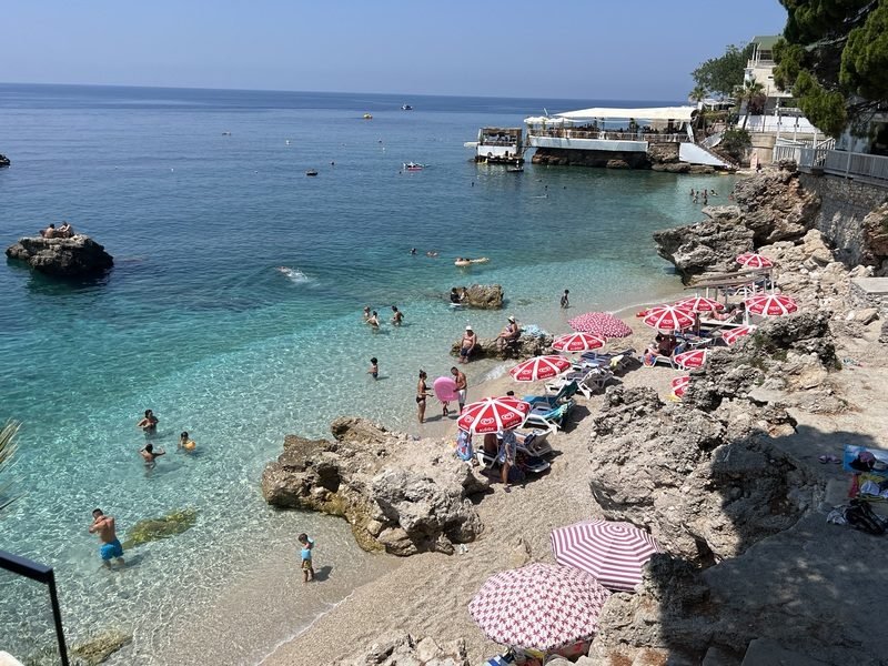 zonnig rotsstrand aan de Albanese Rivièra met helder turquoise water waar mensen zwemmen en ontspannen onder rode en witte parasols bij strandrestaurant